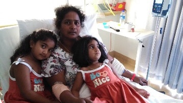 A woman lies in a hospital bed with two children lying on top of her. The girls are dressed in matching red dresses