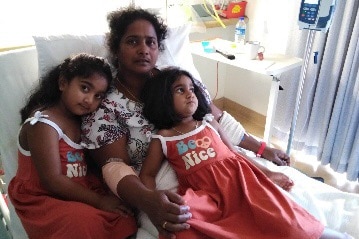 A woman lies in a hospital bed with two children lying on top of her. The girls are dressed in matching red dresses