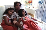A woman lies in a hospital bed with two children lying on top of her. The girls are dressed in matching red dresses