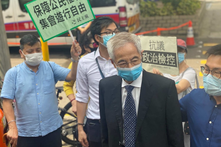 An elderly man in a suit and glasses and a face mask is surrounded by placards in Chinese language.