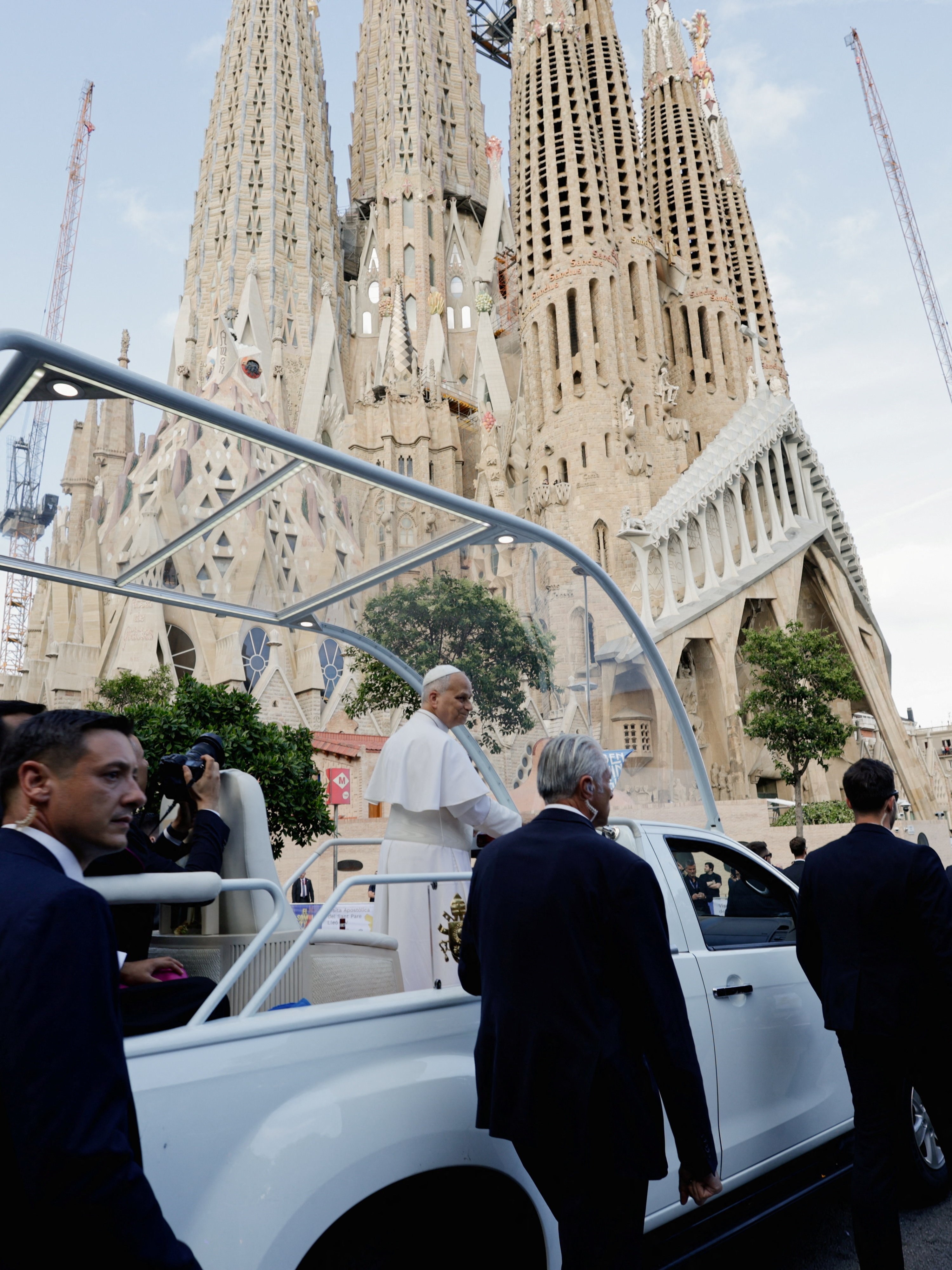 Pope XIV in white robes standing in the back of the popemobile, surrounded by men in suits, in front of the Sagrada Familia.