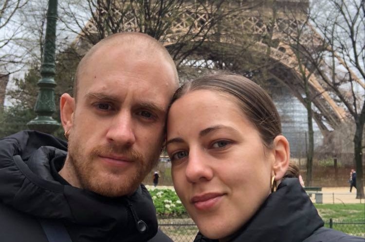 Bridget Caldwell and Jack Bright, a young couple, smile for a selfie in front of the Eiffel Tower.