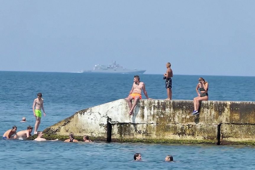 Swimmers enjoy the water at a Crimean beach as a Russian warship passes by in the background