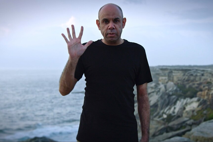 Comedian Steven Oliver standing on a cliff above the ocean, holding up five fingers