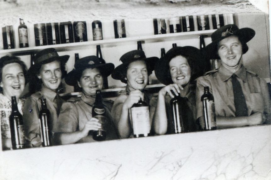 A group of women in uniform, wearing hats, pose for a photo holding big brown bottles