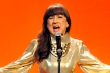 Judith Durham of The Seekers wearing a gold dress and performing with her arms wide in front of an orange background.