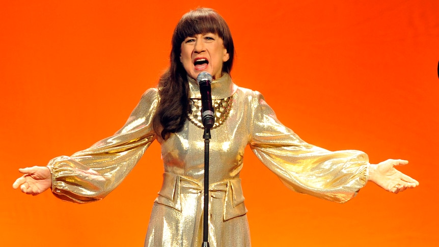 Judith Durham of The Seekers wearing a gold dress and performing with her arms wide in front of an orange background.