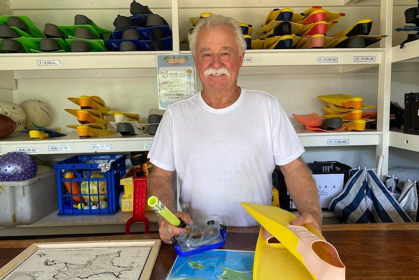 A man with gray hair and a mustache stands behind a counter where snorkels and flippers are sold.