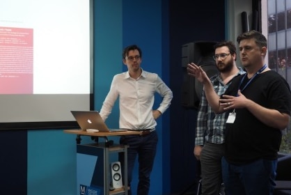 Three men stand at a lecture in front of a screen giving a presentation.