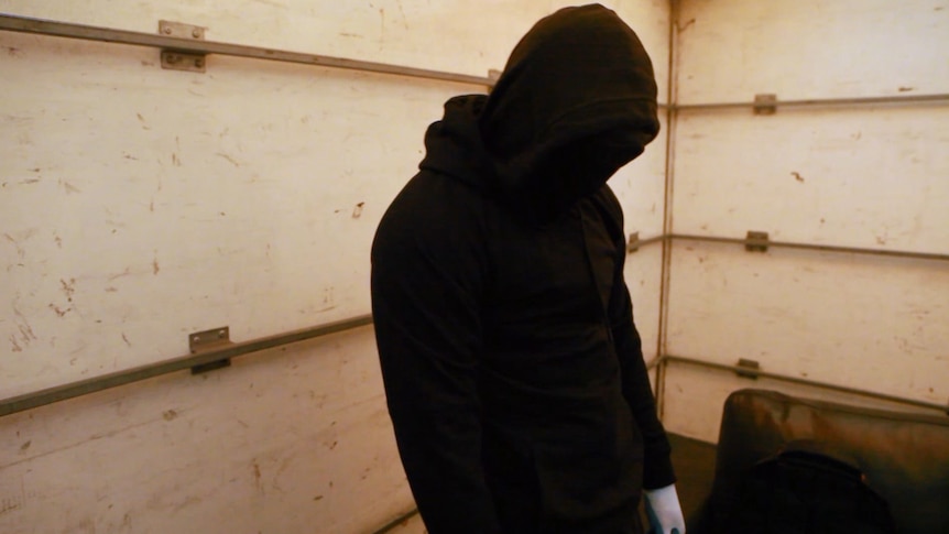 A figure wearing a dark hood, mask and gloves stands in a shipping container with scratched walls.