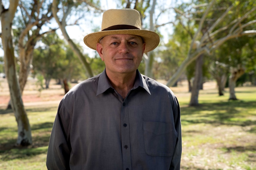 A caucasian man in a dark, long-sleeved shirt and a Panama hat stands outside with trees in the background.