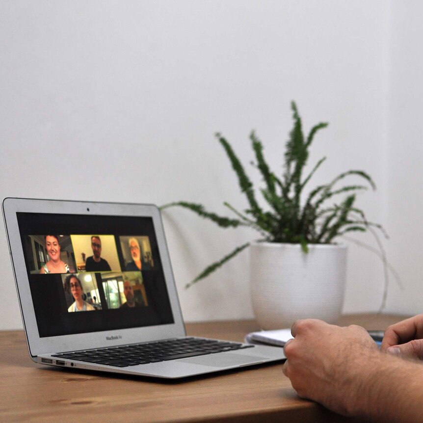 A man sits at a table participating in a Zoom meeting. On the laptop in front of him are five faces of people in the meeting.