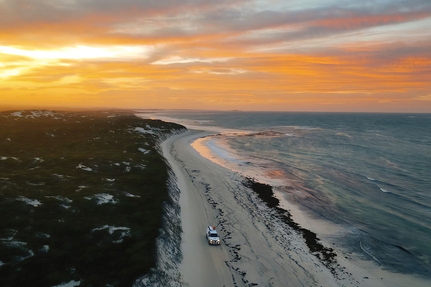 An aerial view of a beach, land, and the ocean. A vehicle is on the beach.