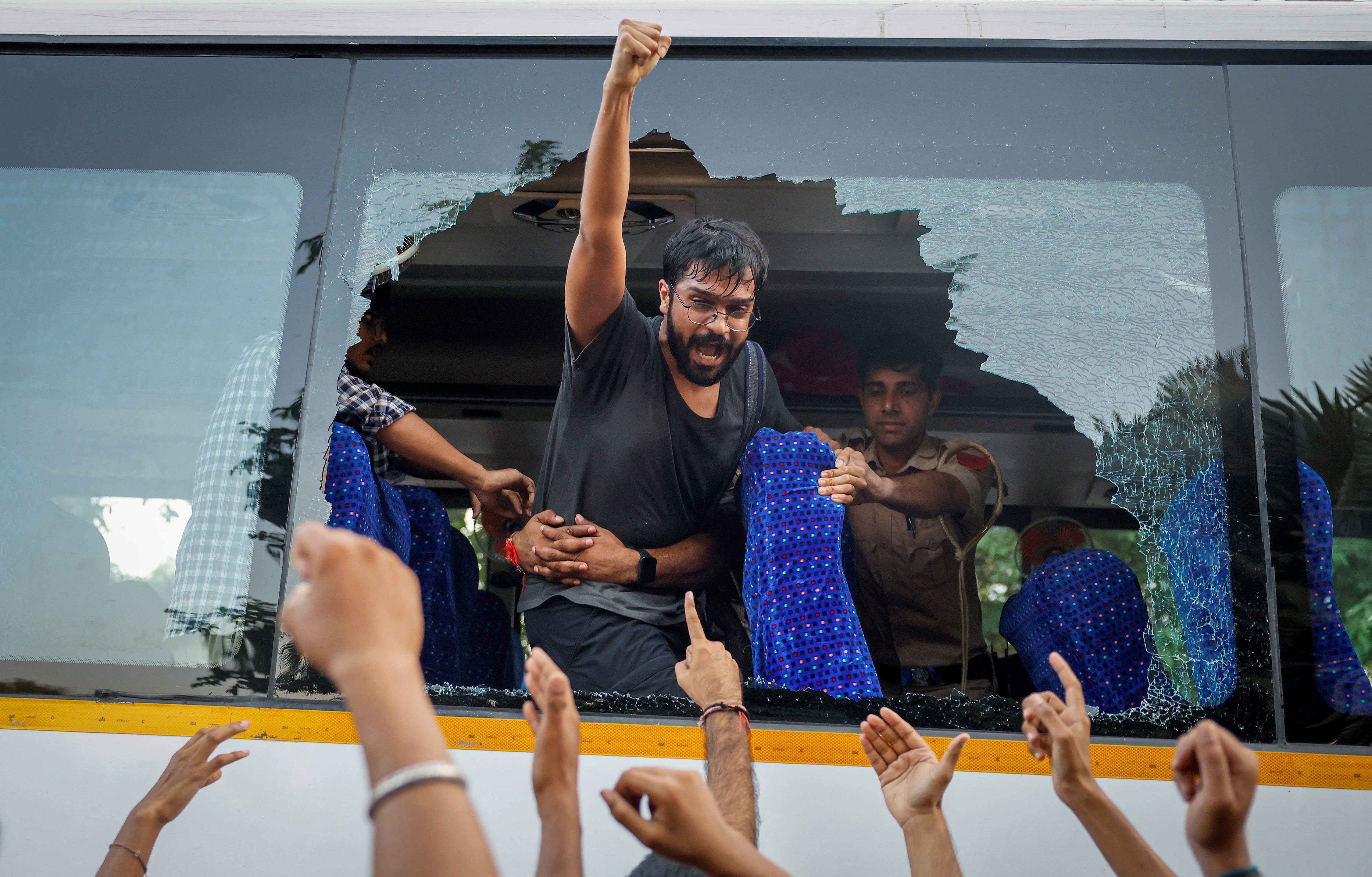 A man raising his fist out a broken bus window at supporters cheering below.