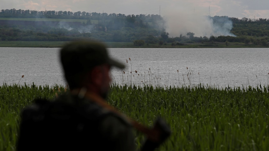 A soldier that is out of focus is in the foreground while in focus in the background is a body of water and smoke. 