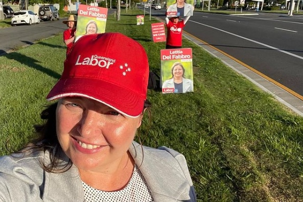 a selfie of a political candidate in a red labor party hat, with two volunteers waving corflutes on the side of a busy rode