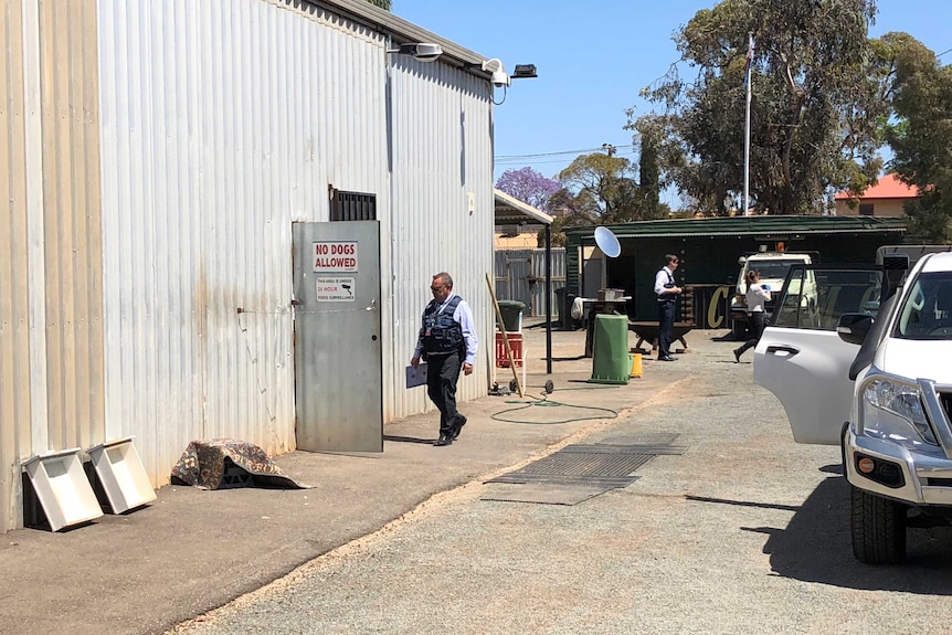 A detective walks towards the door of a shed at the Club Deroes' Kalgoorlie headquarters with other officers and a car nearby.