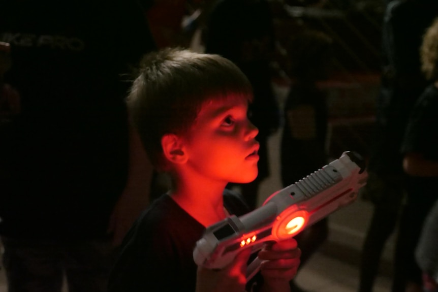 A boy's face is illuminated by the red blow of a laser tag gun he is holding. 