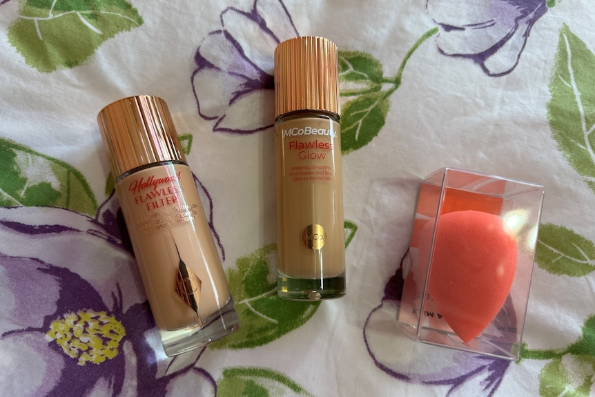 Two circular bottles with gold lids lie flat on a purple floral background. An orange makeup sponge is in the corner.