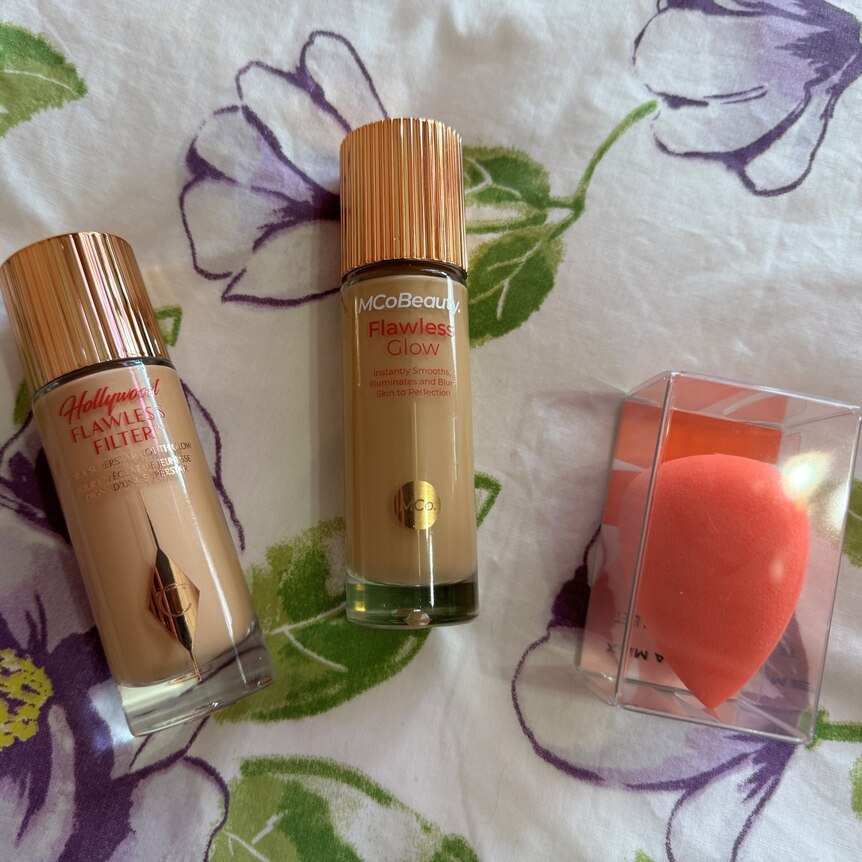Two circular bottles with gold lids lie flat on a purple floral background. An orange makeup sponge is in the corner.