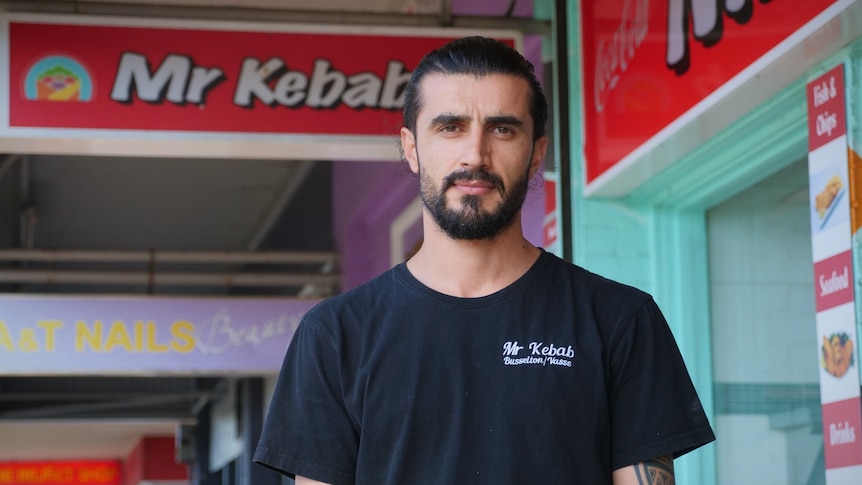A serious young man, black hair slicked back, beard, wears black t-shirt, in front of a Mr Kebab shop sign.