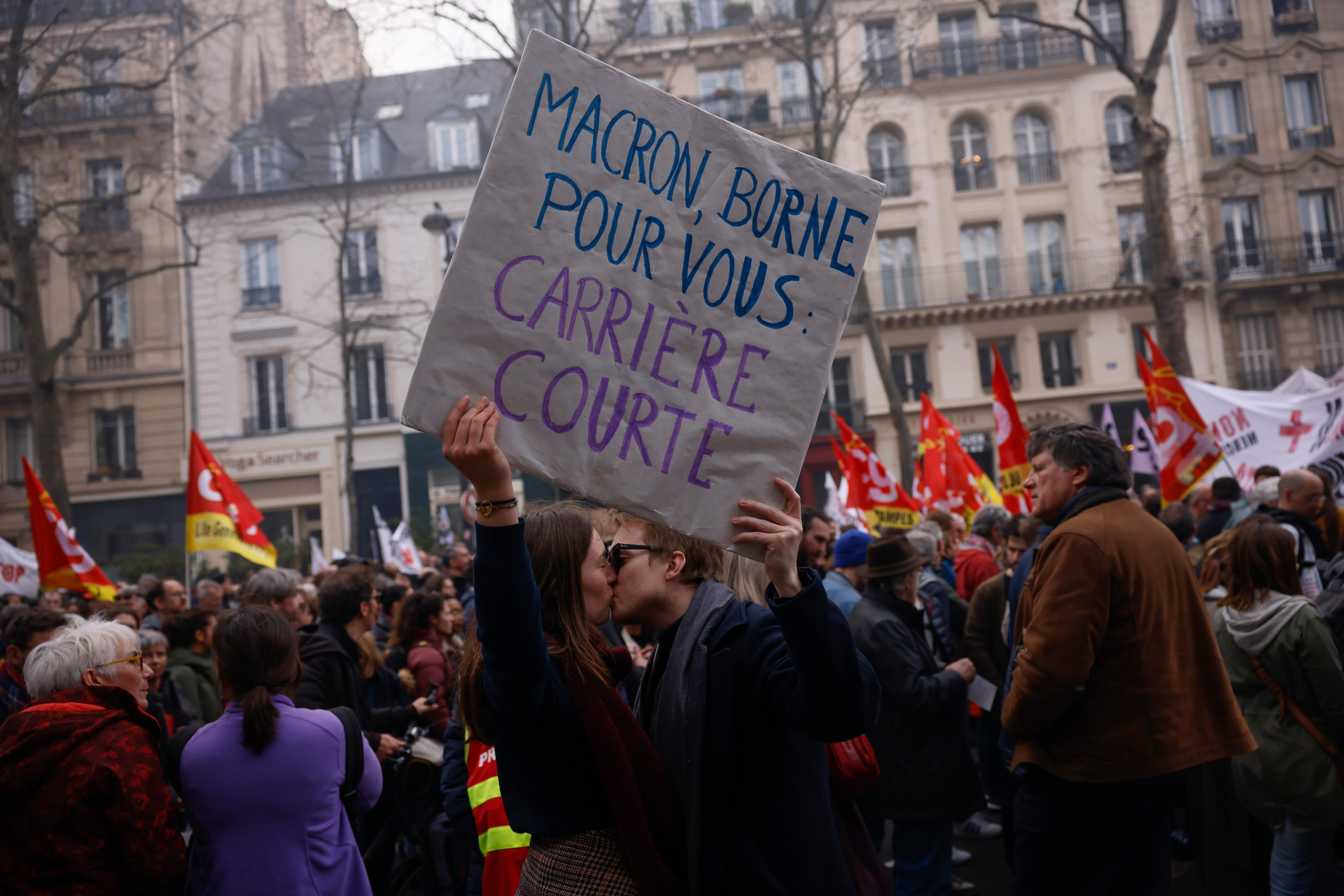 two protesters kiss as they hold up a sign which reads in French: "Macron Borne for you a short carrier" 