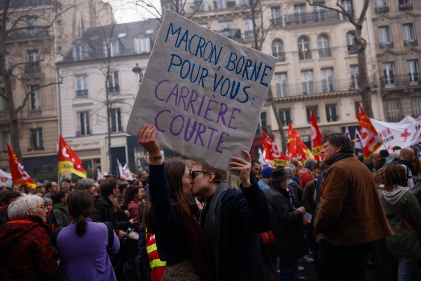 two protesters kiss as they hold up a sign which reads in French: "Macron Borne for you a short carrier" 