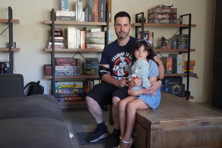 Man sitting on a coffee table in front of bookcases with board games, holding his young daughter.