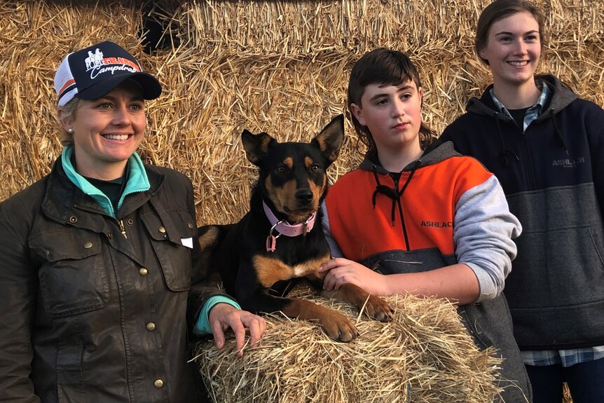 Two young women and a young man are grinning next to a black and tan kelpie sitting on stacked up hay bales. 