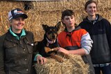 Two young women and a young man are grinning next to a black and tan kelpie sitting on stacked up hay bales. 