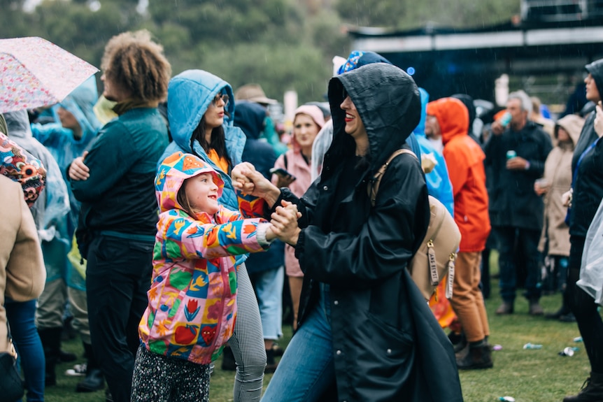A woman in a black raincoat and child in a colourful raincoat dance together in a fiel
