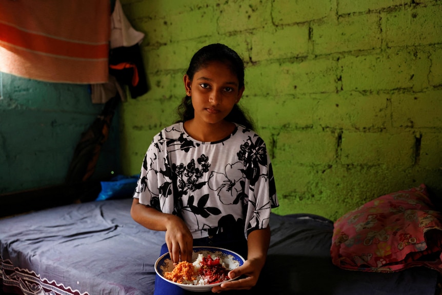 A teenage Sri Lankan girl uses her hand to eat food off a plate while sitting on her bed and looking at the camera.
