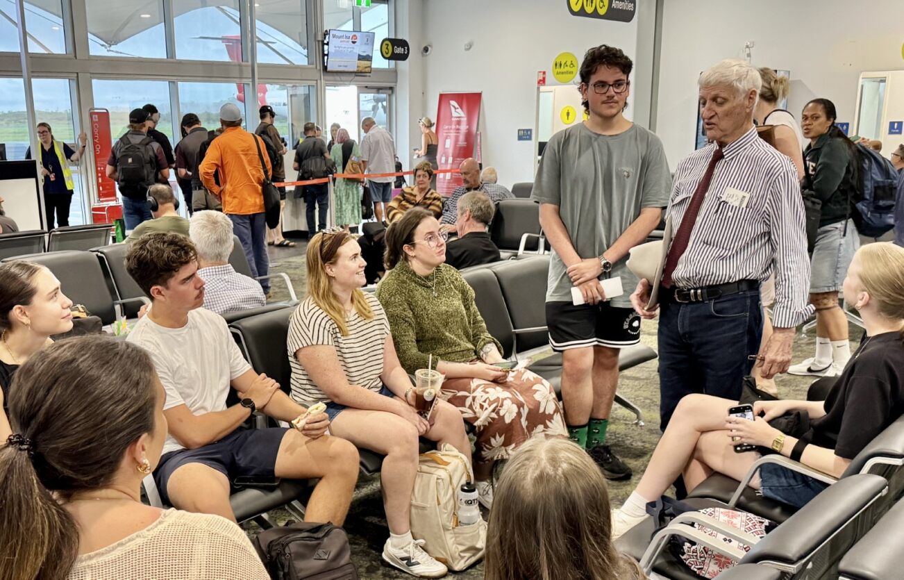 El representante Bob Katter se encuentra en un aeropuerto y habla casualmente con un grupo de estudiantes sentados. 