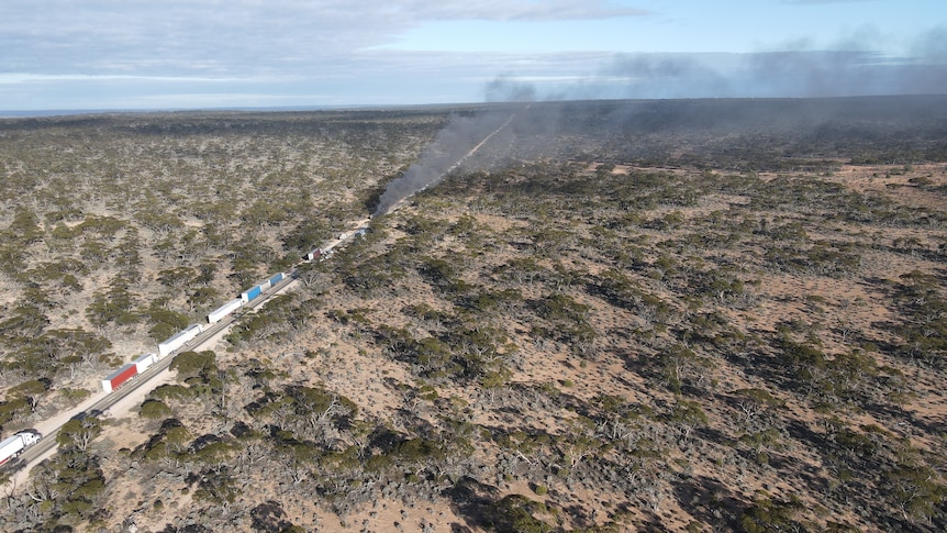 A vehicle burning and emitting black smoke as trucks were banked up on a road among scrubland