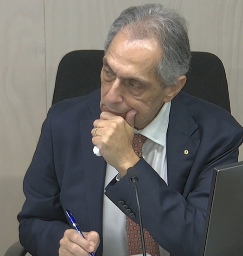 John strokes his chin and has a look of concentration. He has grey hair and a dark blazer.