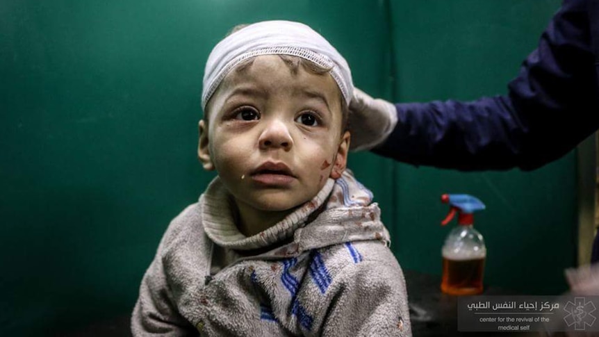 A toddler with short brown hair with a bandage around his head, and drops of blood on his face, shirt and jumper
