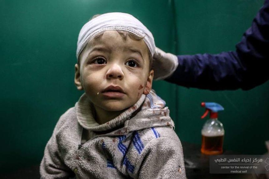 A toddler with short brown hair with a bandage around his head, and drops of blood on his face, shirt and jumper