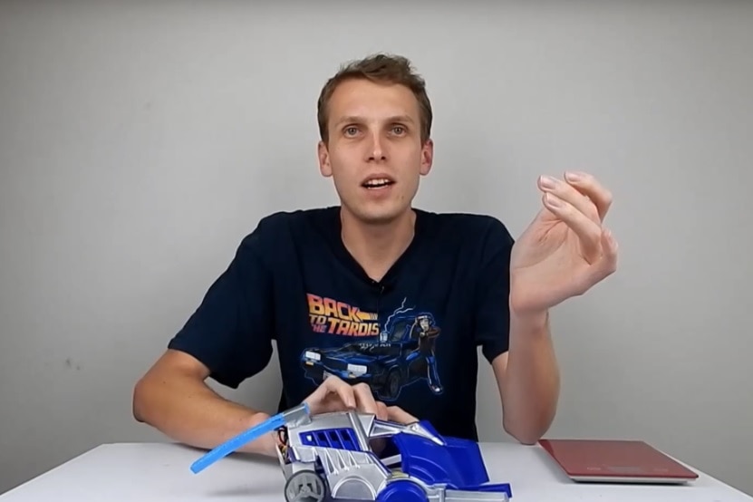 A man sits at a desk with a robot in his hand as he talks.