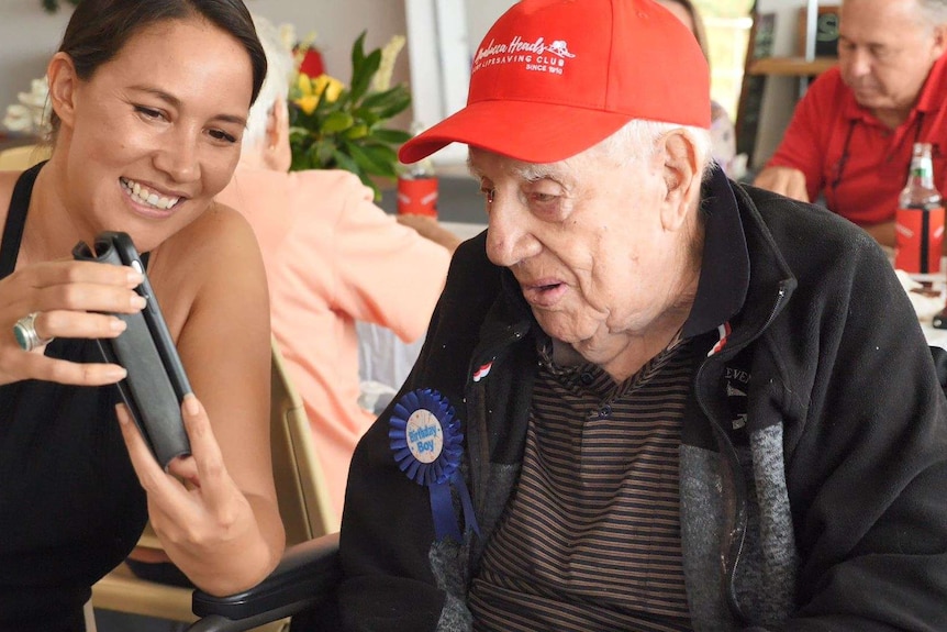 An elderly man in a red baseball cap facetiming on a phone held up by a grinning young woman next to him.