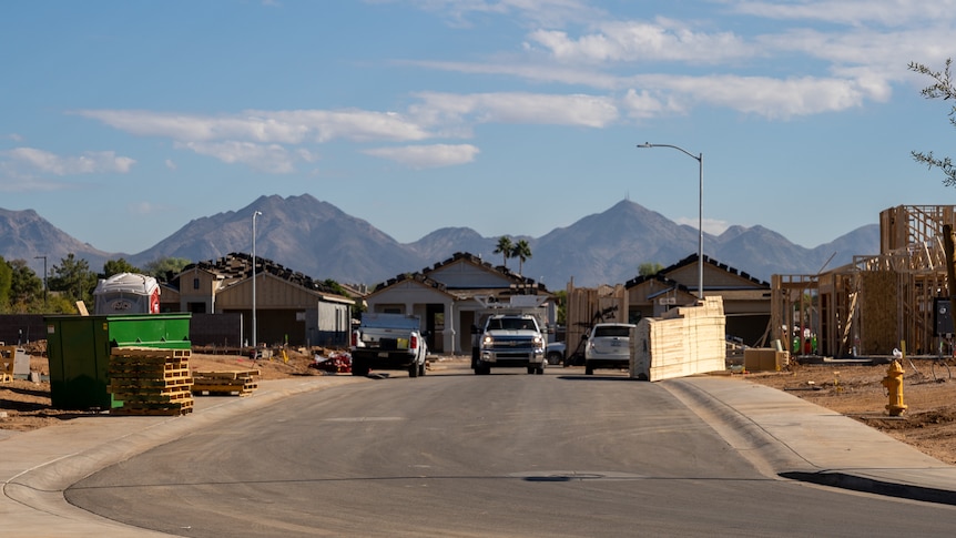 A few utes are parked at the end of a street with houses under construction at the end and a skip bin on one side.