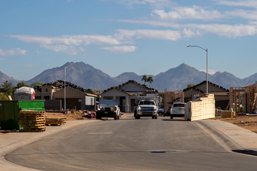 A few utes are parked at the end of a street with houses under construction at the end and a skip bin on one side.