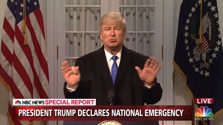Alec Baldwin wears a grey-orange wig, fake tan and a suit while standing at a podium in front of US flags.