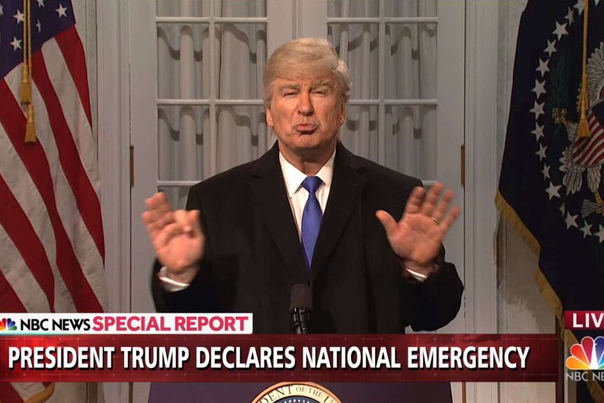 Alec Baldwin wears a grey-orange wig, fake tan and a suit while standing at a podium in front of US flags.