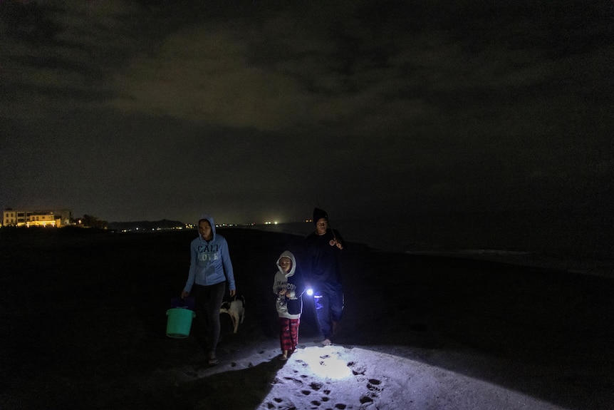Three people walk on the sand in the dark holding torches and buckets. 