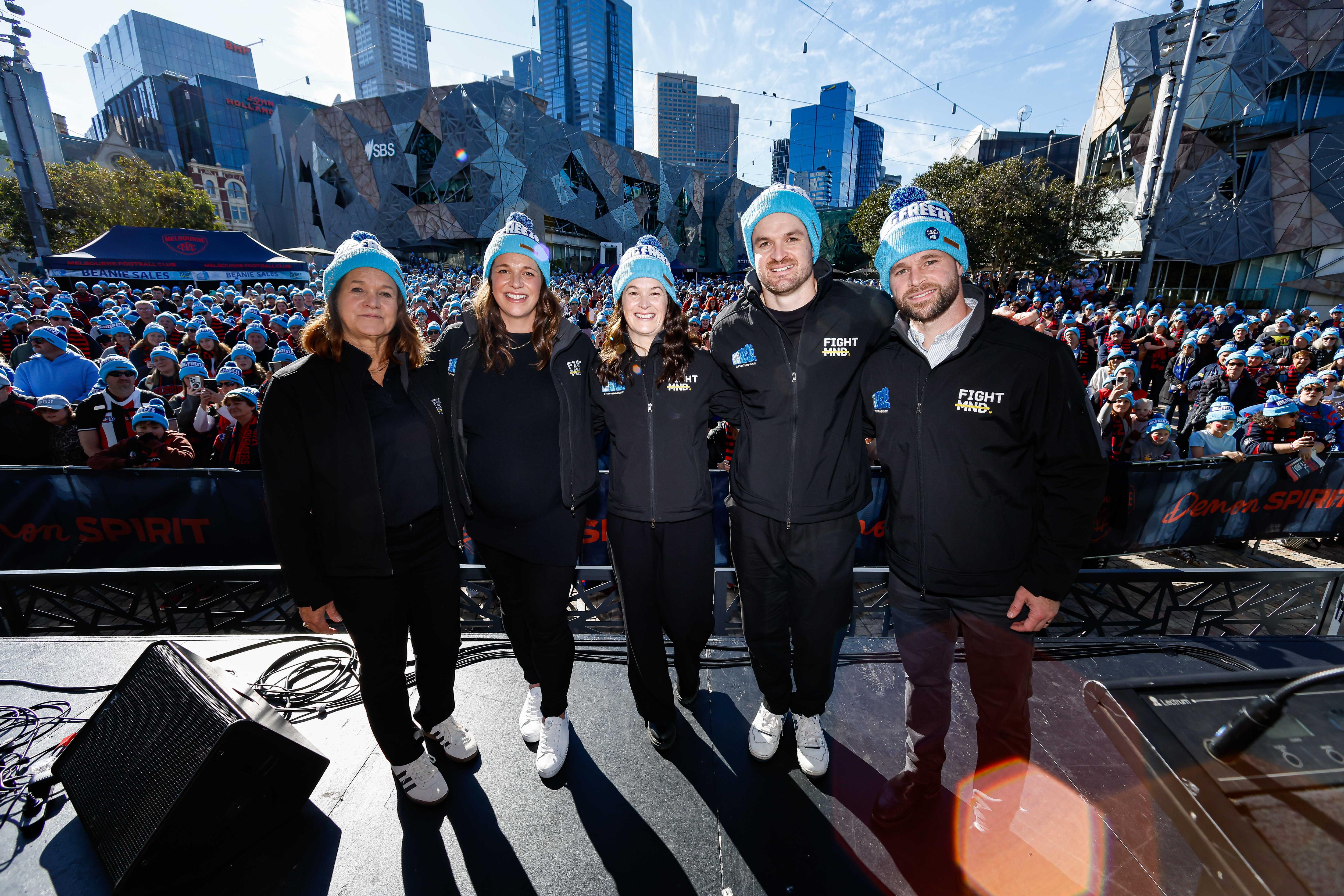 Three women and two men dressed all in black with blue beanies stand on a stand in front of a large crowd with the same beanies.