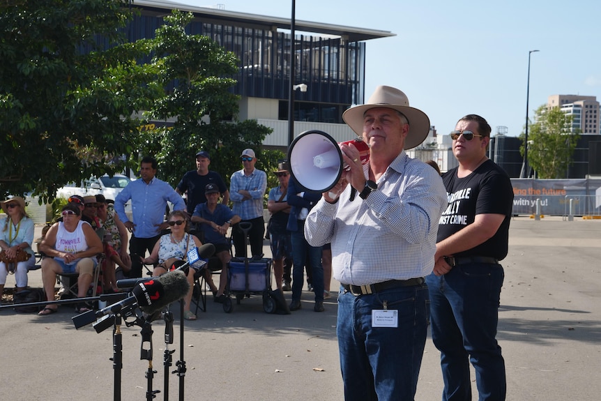 A man in a button down shirt and cowboy hat speaks into a megaphone surrounded by protestors. 