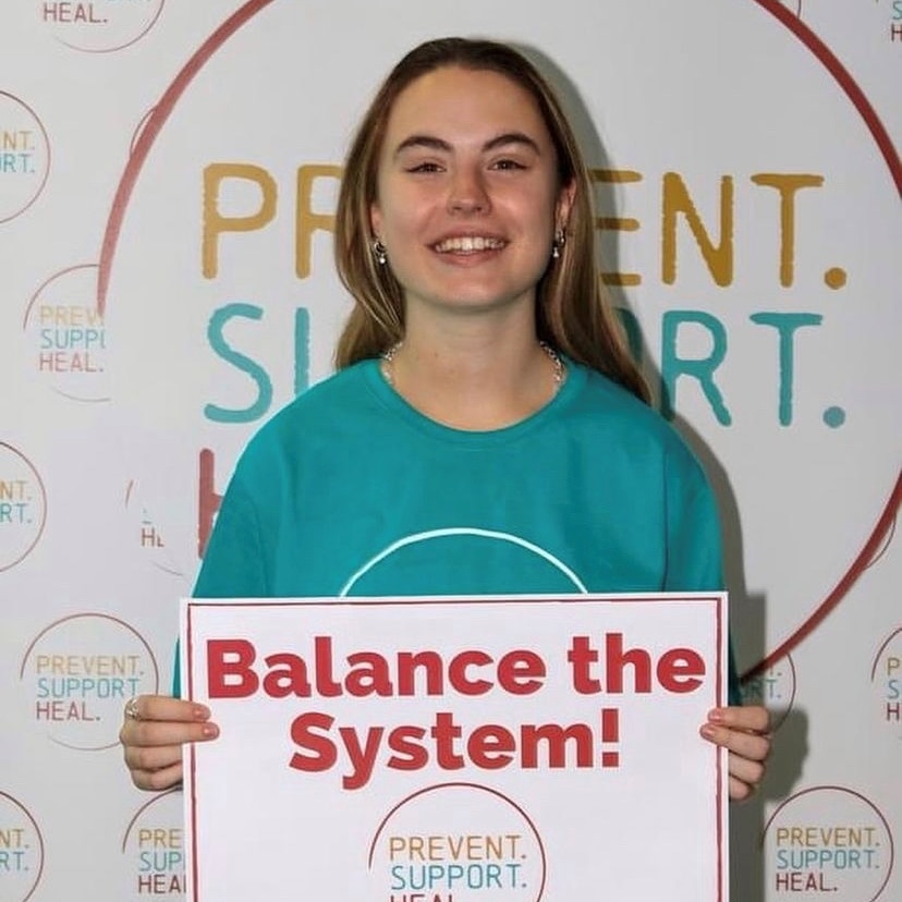 A young woman wearing a teal T-shirt holds up a sign that says 'Balance the system!'