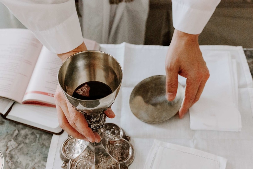 The hands of a priest reach for red wine in a metal goblet