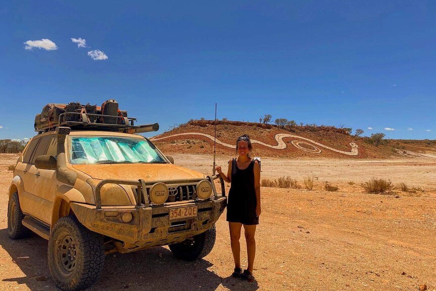 A woman stands next to a mud-covered 4WD in the outback in front of a huge Indigenous serpent mural on a sand dune.