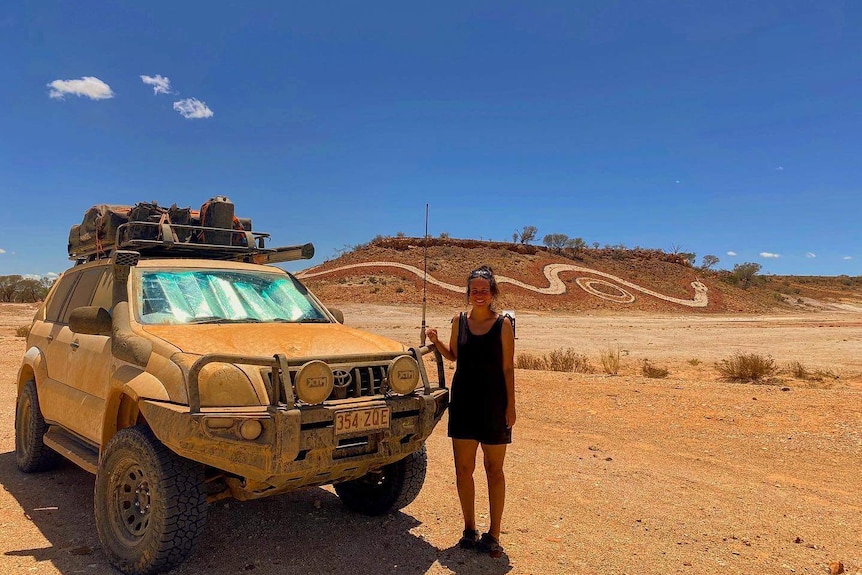 A woman stands next to a mud-covered 4WD in the outback in front of a huge Indigenous serpent mural on a sand dune.
